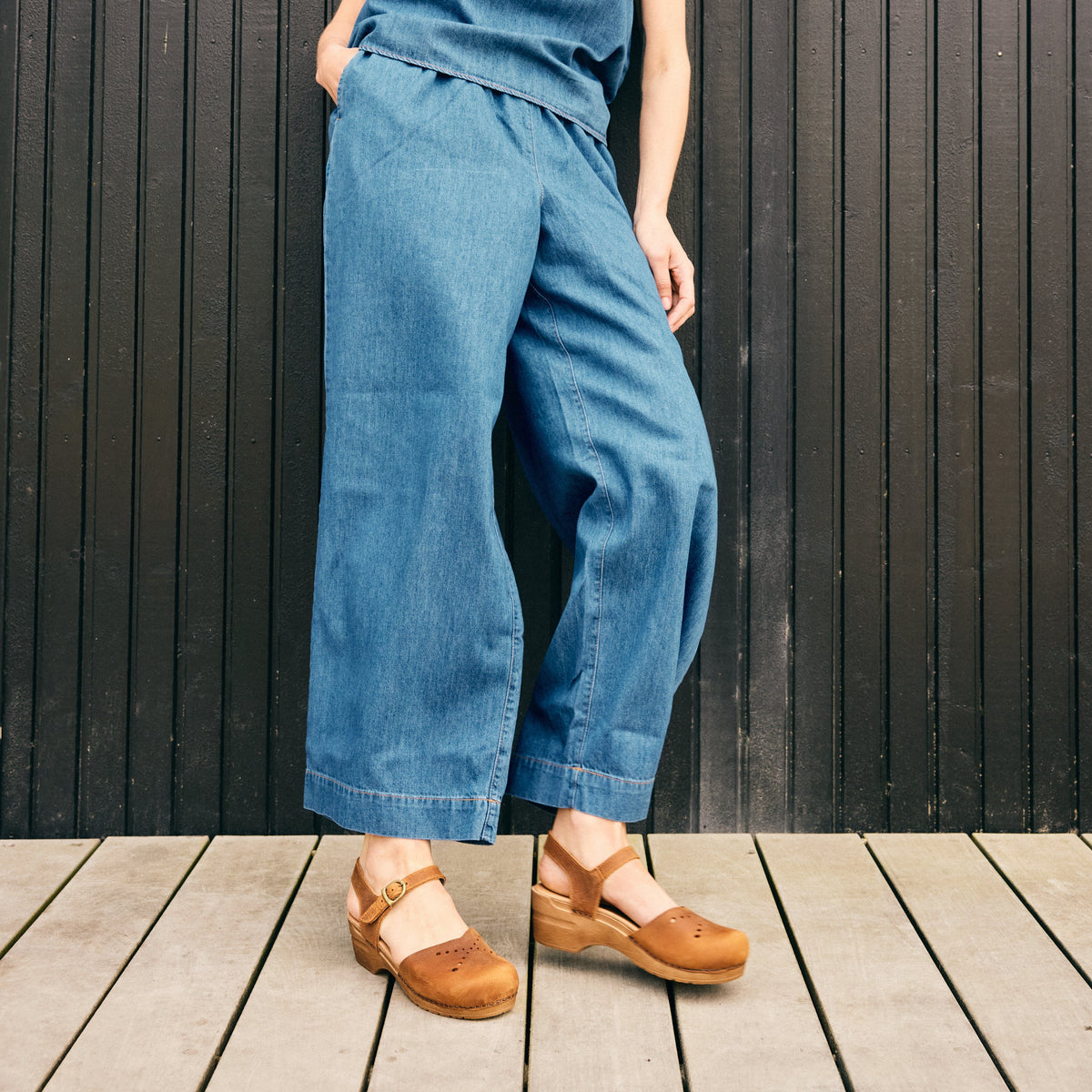 Woman wearing denim outfit against black wall with chestnut colored sandals