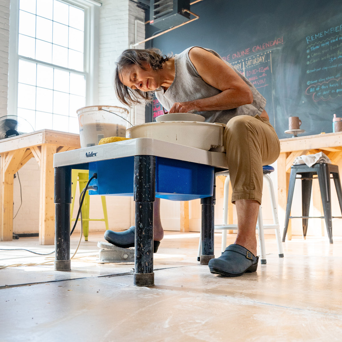 Older woman working on pottery wheel wearing blue clogs.