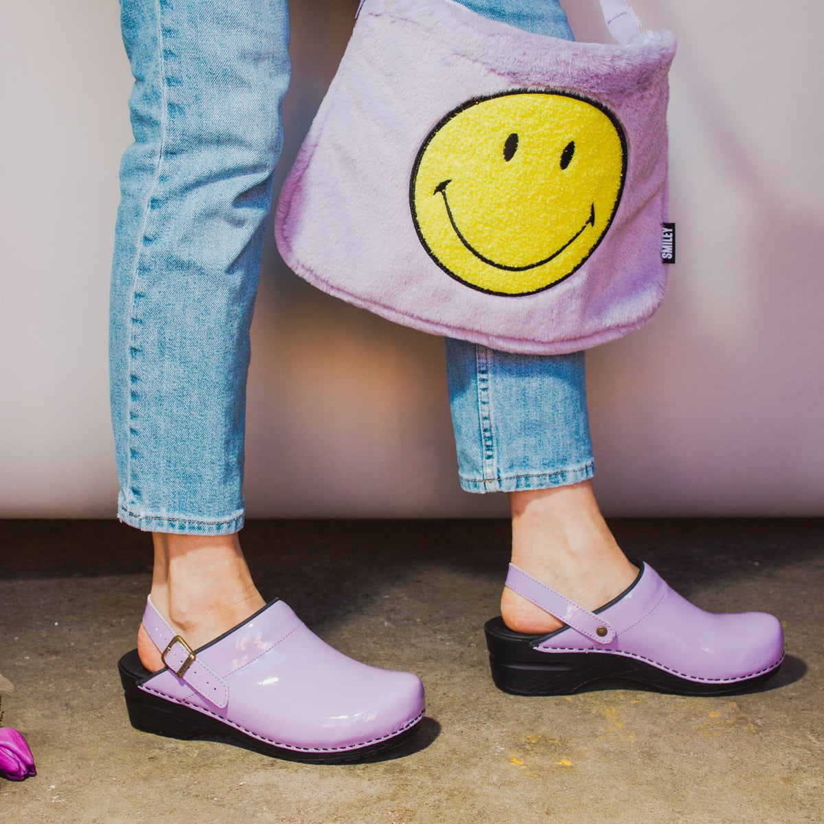 Person wearing jeans and lilac colored slingback clogs carrying a fuzzy lilac messenger bag with a big yellow smiley face on it.