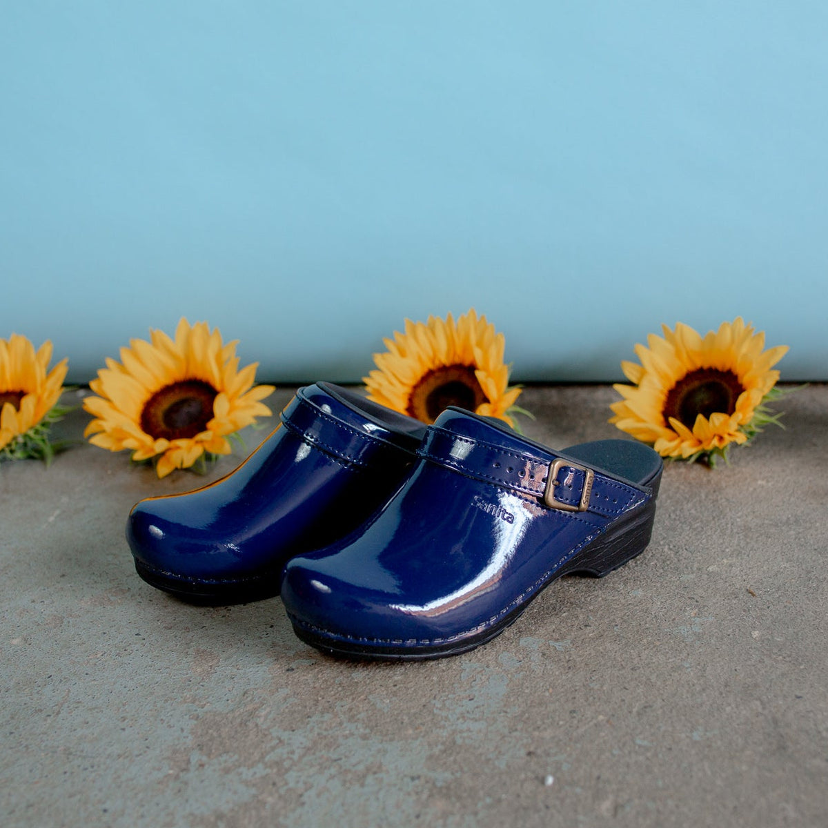 Patent blue shoe on cement floor with light blue background with sunflower heads behind shoe.