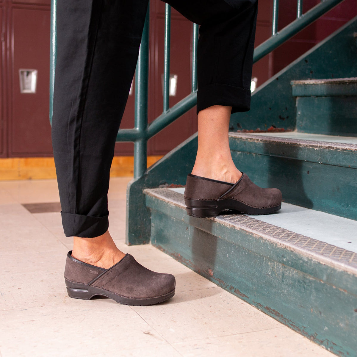Person wearing black cuffed pants with one leg on a step in a school setting. They are wearing steel-colored suede clog shoes.